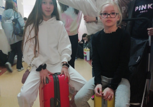 Z lewej strony dziewczynka ubrana na biało siedzi na czerwonej walizce. Obok niej na żółtej walizce siedzi koleżanka z klasy. Za nimi stoją cztery dziewczynki.
